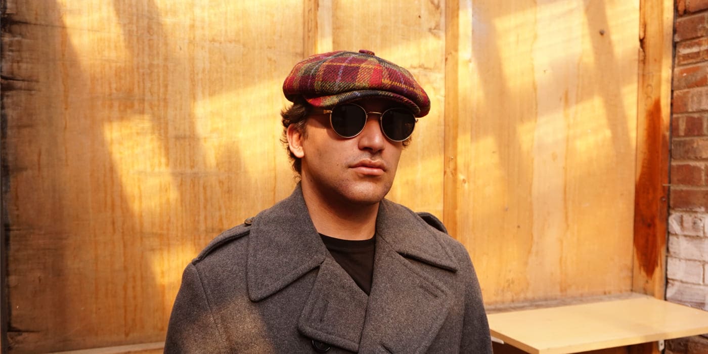 Man wearing a plaid cap and sunglasses against a wooden wall.