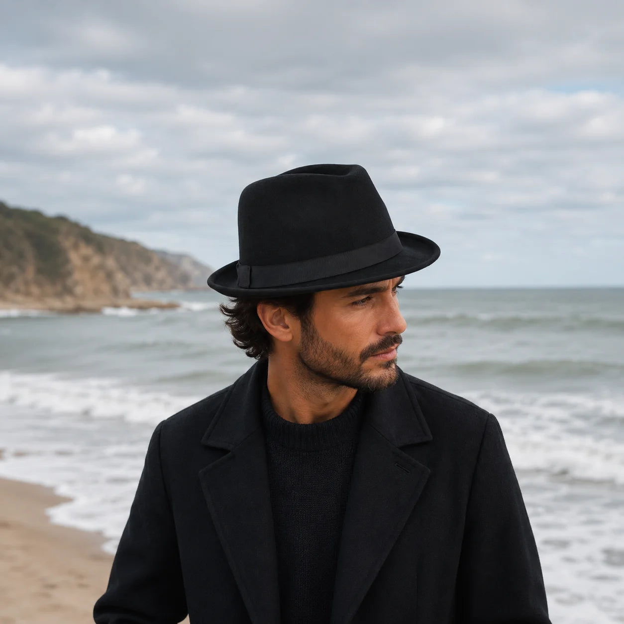 Man wearing a black hat and coat standing on a beach with ocean and sky in the background